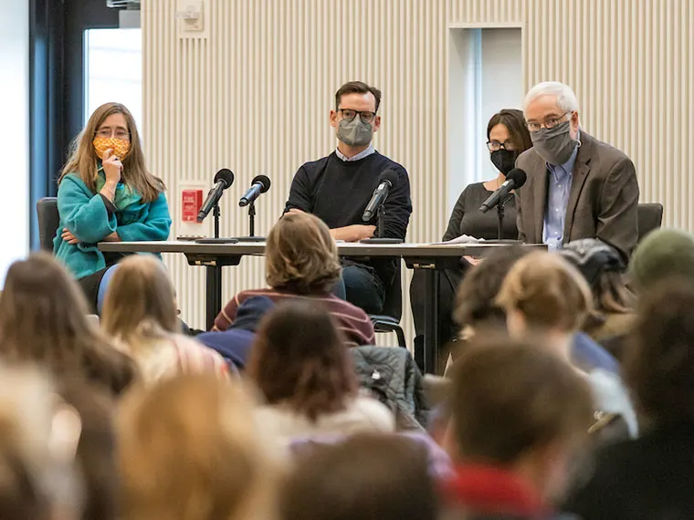 Four individuals wearing masks sit at a panel table with microphones, facing an audience in a conference room.