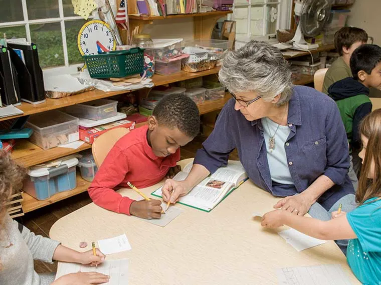 Person assisting a child writing at a round table in a classroom, with books and educational supplies on shelves. Other children are nearby.