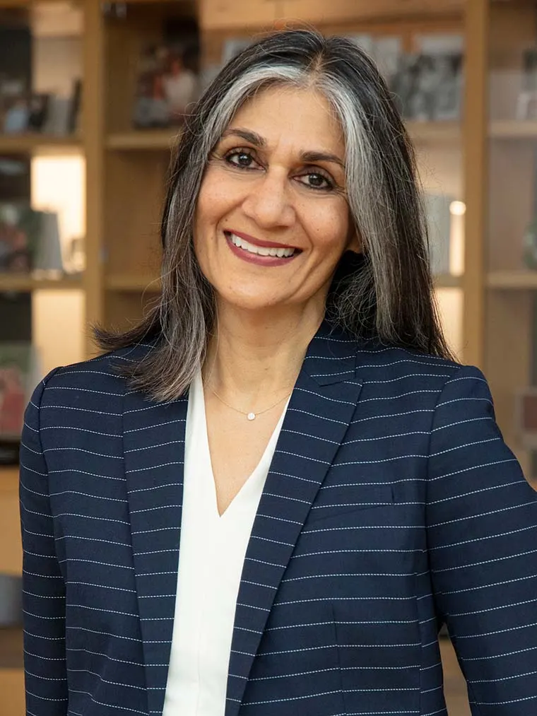 Person with long gray hair smiles, wearing a dark pinstripe blazer and white top, standing against a blurred bookshelf background.