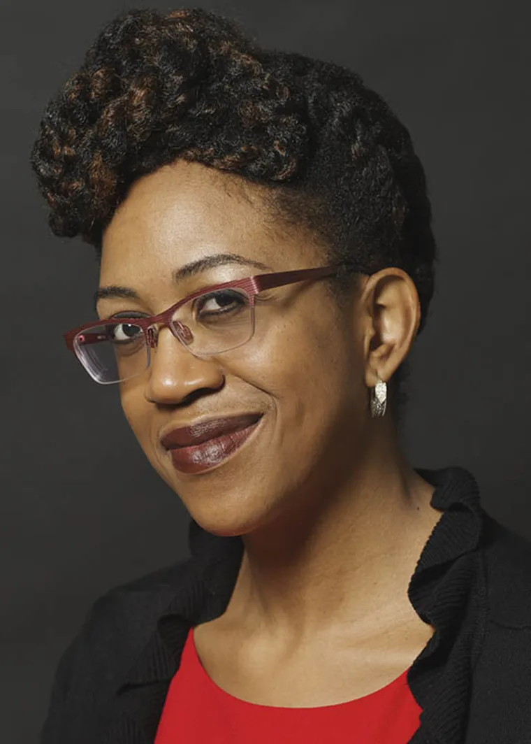 Person with braided hair, wearing red glasses and a red top, smiles slightly against a dark background.