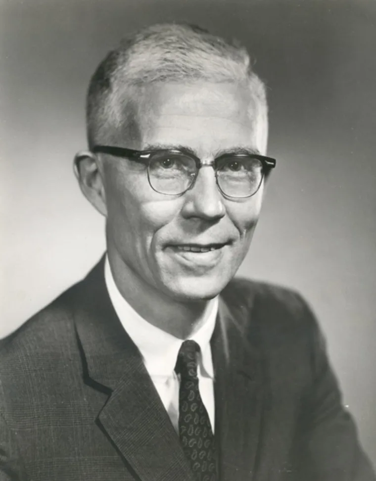 Person with short light hair, wearing glasses, a suit, and tie, looking forward with a neutral expression in a black-and-white photo.