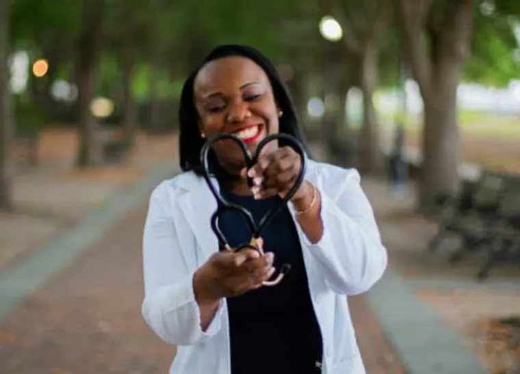 Person in a white coat smiling, holding a stethoscope on a tree-lined pathway.