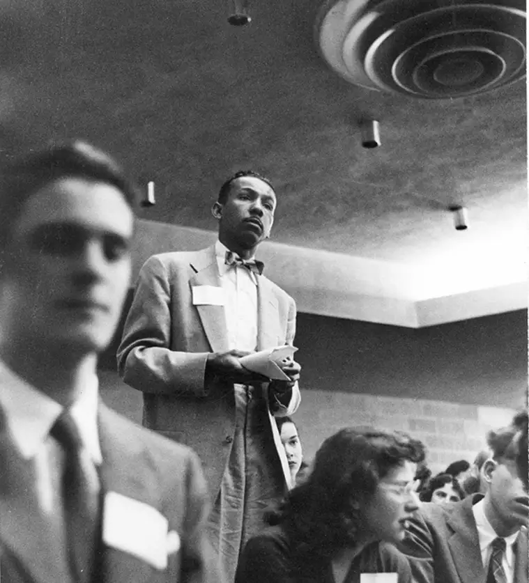 Person in a suit stands in a crowded room, holding papers, appearing to speak or present. Others sit and listen attentively. Ceiling lights are visible.