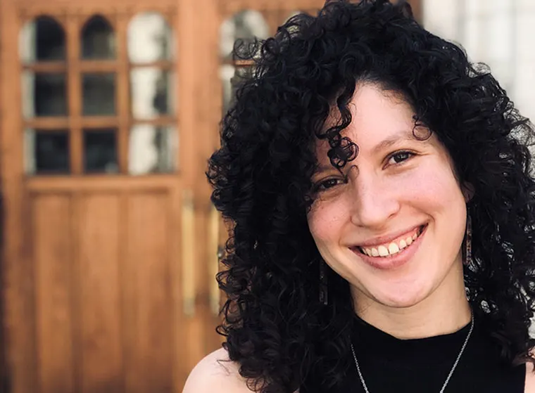 Person with curly hair smiling in front of a wooden door with arched windows.