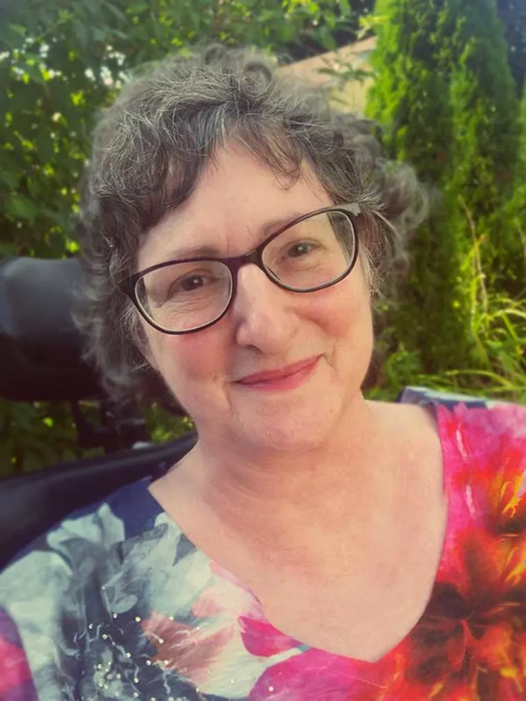Person with glasses and short hair smiles, wearing a colorful floral shirt. Background is a garden with greenery.