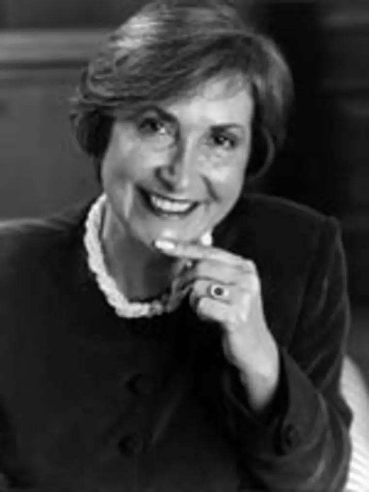 Smiling individual with short hair, wearing a dark outfit and a beaded necklace, resting chin on hand, in a black and white photo.