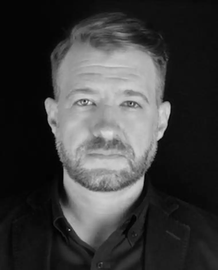 Person with short hair and beard, wearing a collared shirt and jacket, against a dark background.
