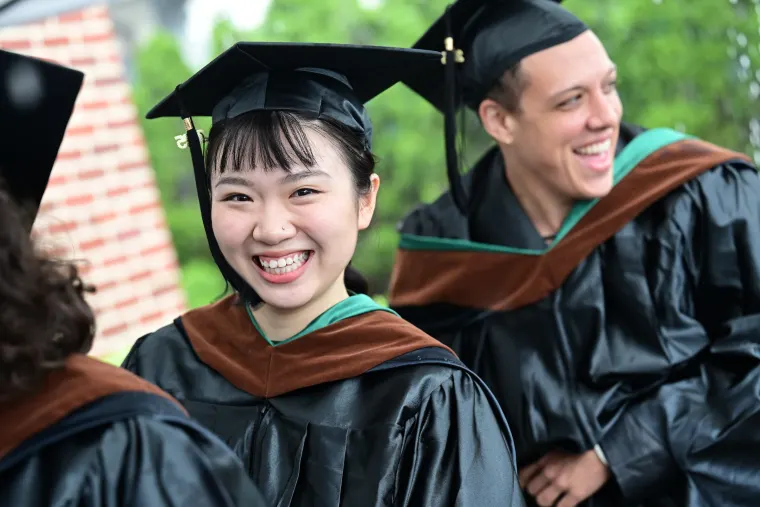 Graduates in black gowns and caps smiling outdoors, with one focused in the foreground, others in the background.