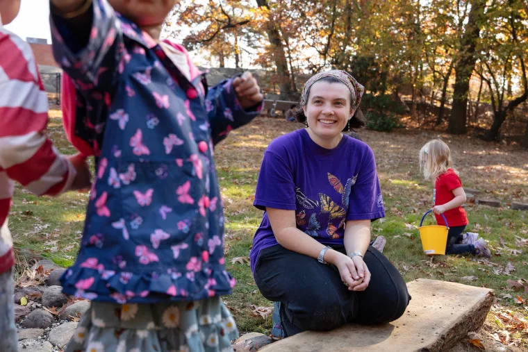 Person kneeling and smiling outdoors with children playing. Child in butterfly jacket foreground; another with yellow bucket in the background.