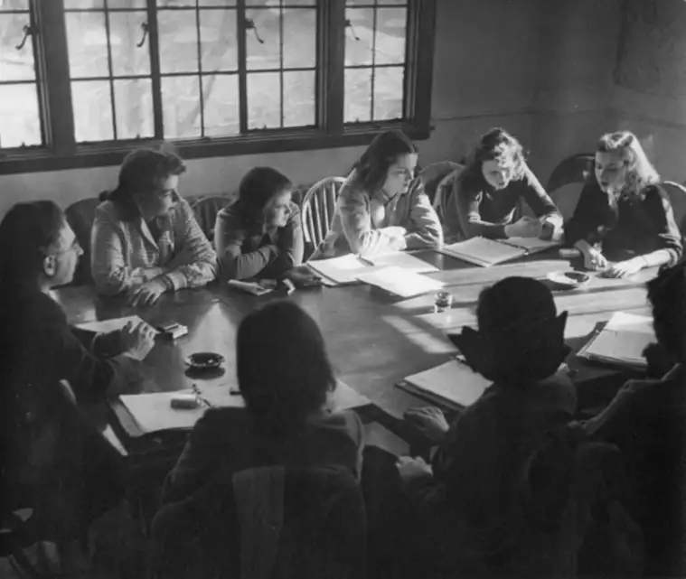 A group of people sit around a table with books and papers, engaged in a discussion in a room with large windows.