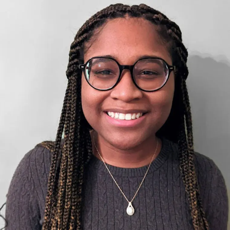 Smiling person wearing glasses and braids, in a dark sweater with a pendant necklace.