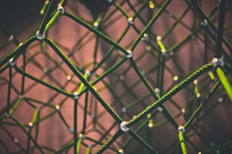 A close-up of a complex structure of intertwined green ropes connected at white junctions, forming a geometric pattern against a blurred background.