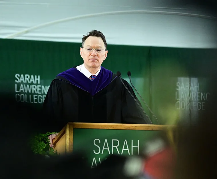 A speaker in academic regalia stands at a podium with microphones, in front of a green backdrop with "Sarah Lawrence College" written on it.