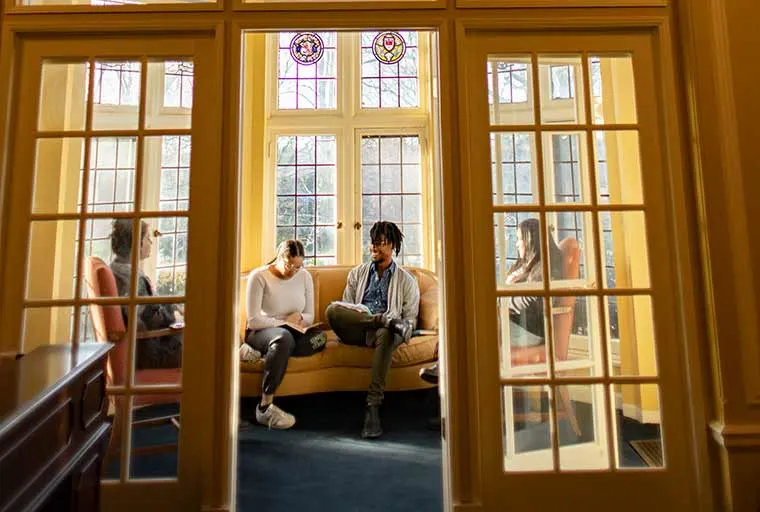 Four people sit together in a sunlit room with large windows, engaged in conversation and reading, surrounded by stained glass and wooden furniture.