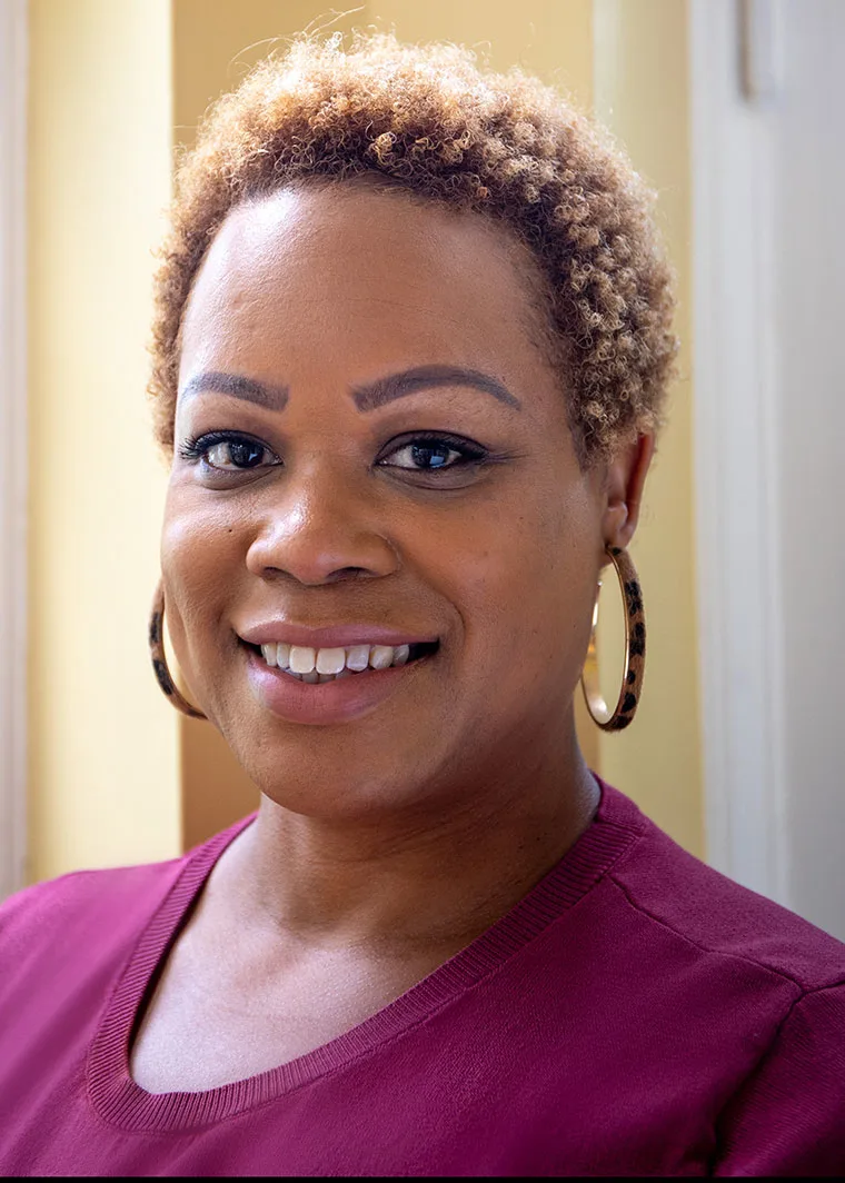 Smiling person with short curly hair and hoop earrings, wearing a maroon top, stands in a sunlit room.