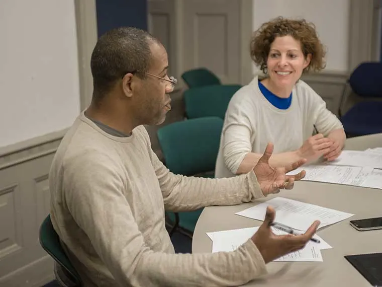Two people are seated at a table with papers, engaged in a conversation. One is gesturing while speaking, and the other is smiling.