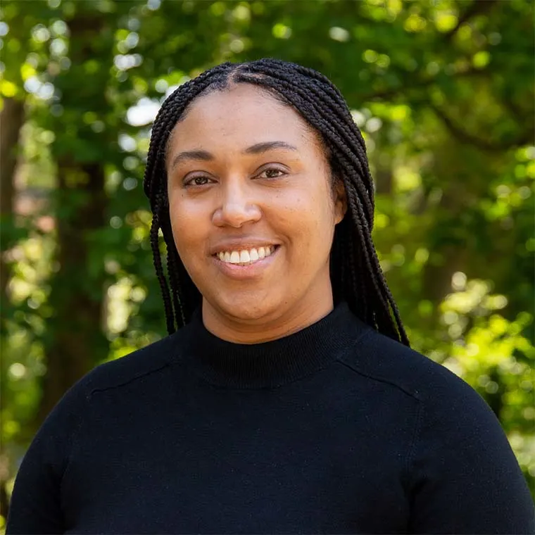 Person with braided hair smiling in a black sweater, standing outdoors with trees in the background.