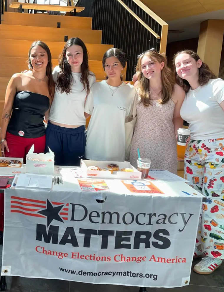 Five people stand behind a table with a "Democracy Matters" banner, smiling, with donuts and drinks on the table.