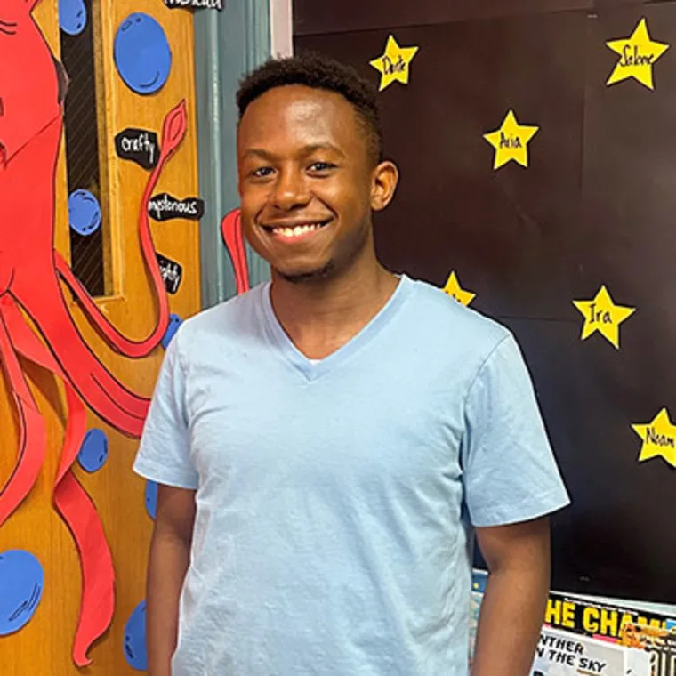 Person smiling in a classroom with colorful decorations and star-shaped name tags on the wall.