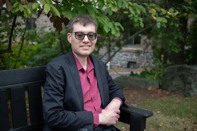 Person in sunglasses sitting on a bench outdoors, wearing a dark jacket and red shirt, with greenery and a brick building in the background.