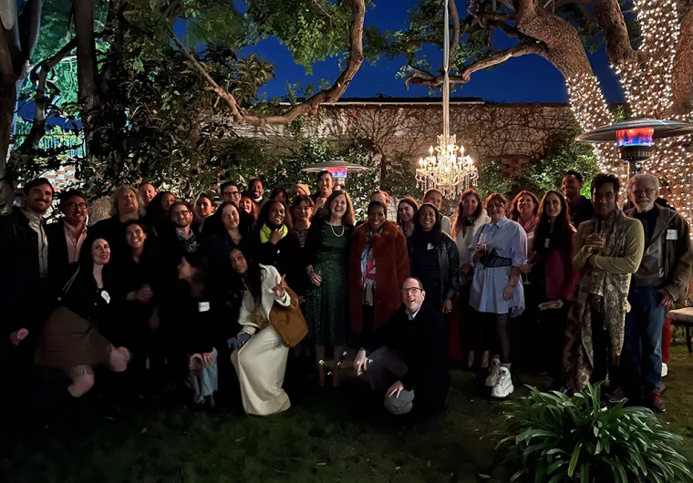 A large group of people smiling outdoors at night, with string lights in trees and a chandelier illuminating the background.