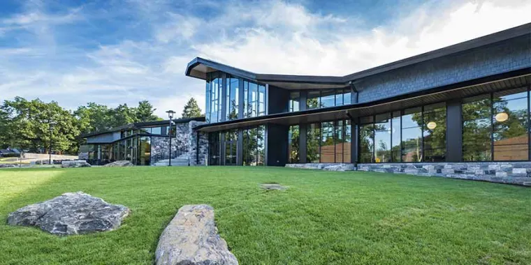 Modern building with large glass windows, surrounded by a green lawn and rocks, set against a sky with clouds and trees in the background.