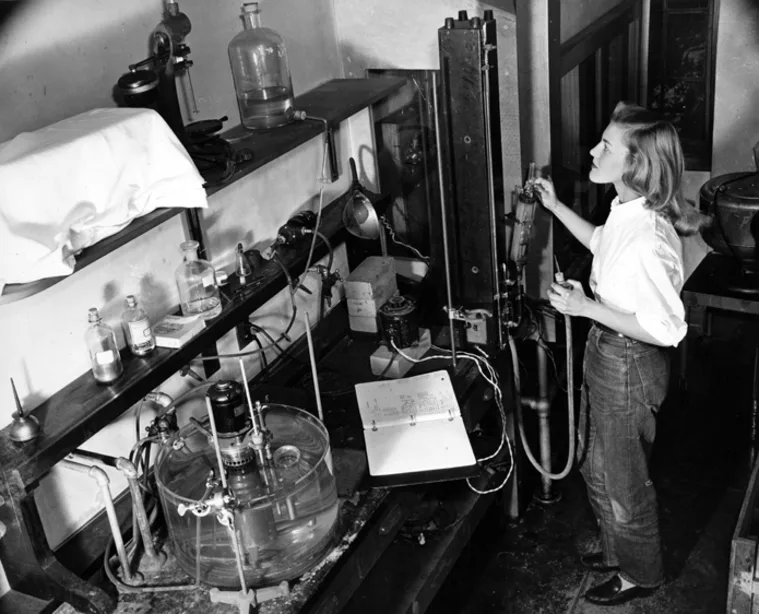 Person in a lab working with scientific equipment, including a large device and various bottles on shelves.