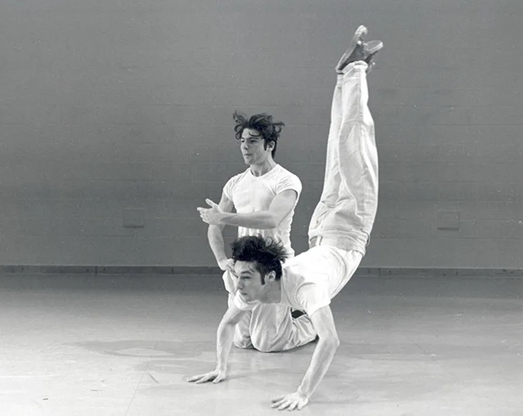 Two dancers in white clothing perform a contemporary dance. One dancer balances on their hands, legs in the air, while the other gestures nearby.