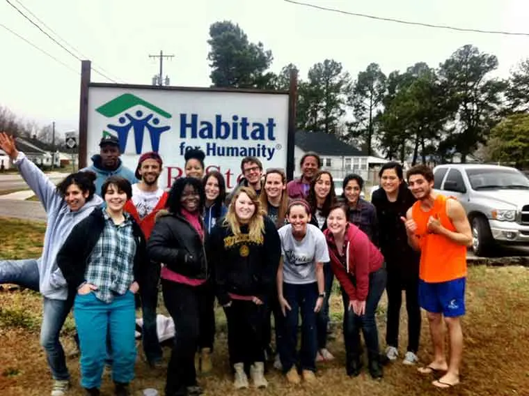 A group of people stands smiling in front of a Habitat for Humanity sign, in an outdoor setting with trees and a parked truck nearby.