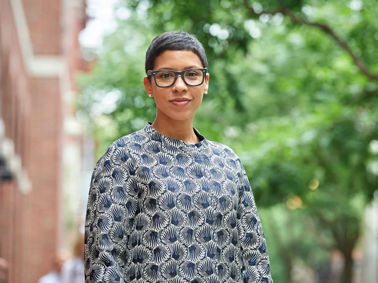 Person with short hair and glasses wearing a patterned top stands on a tree-lined street.