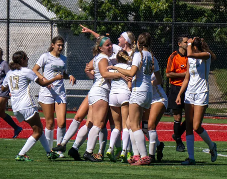 Soccer team in white uniforms celebrating on the field, with a referee nearby in an orange shirt.