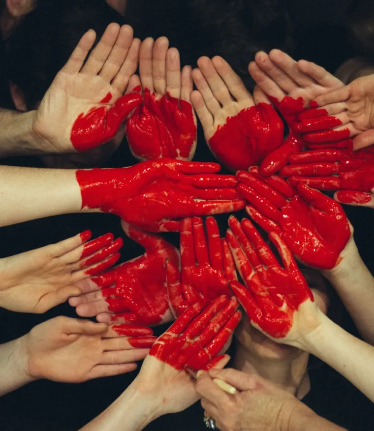 Multiple hands covered in red paint forming a heart shape on a dark background.