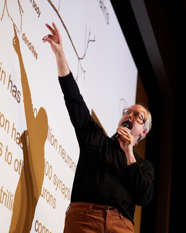 A person holding a microphone points towards a diagram of neurons on a large screen during a presentation.