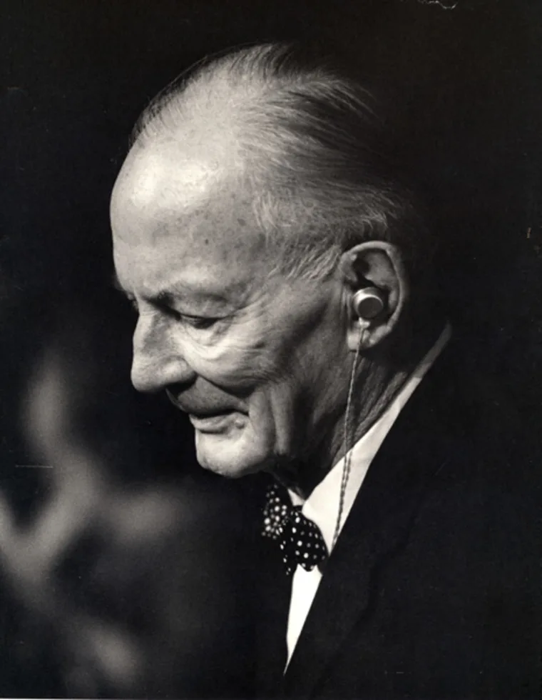 Elderly person with a hearing aid and bow tie, shown in profile in a black and white photo.