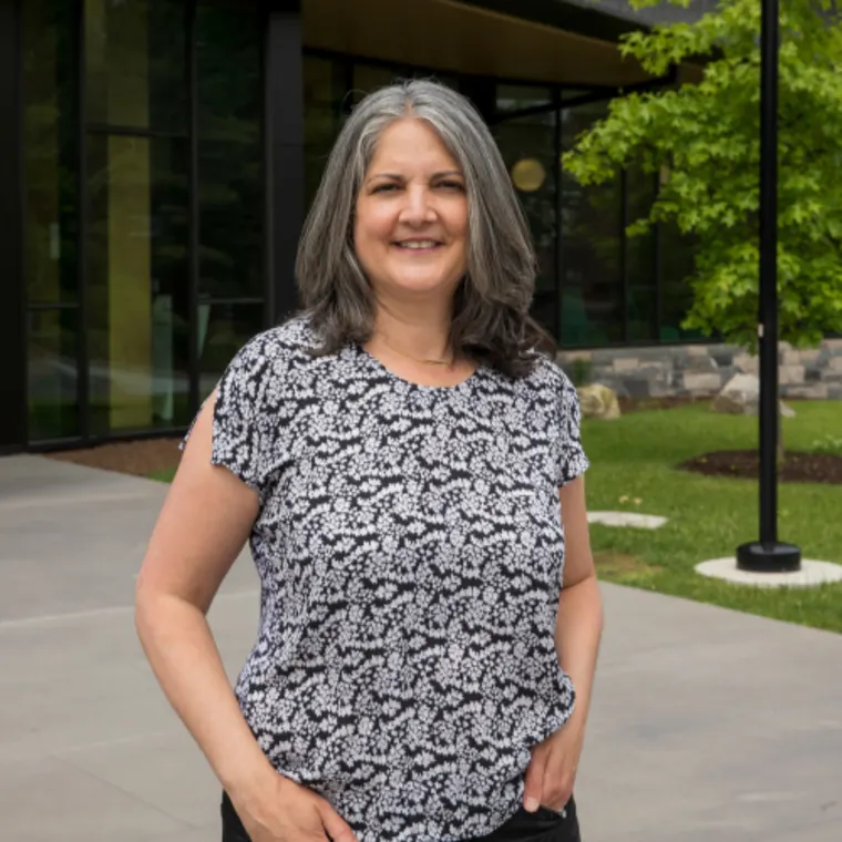 Person with gray hair stands outside a modern building, wearing a patterned shirt and smiling.
