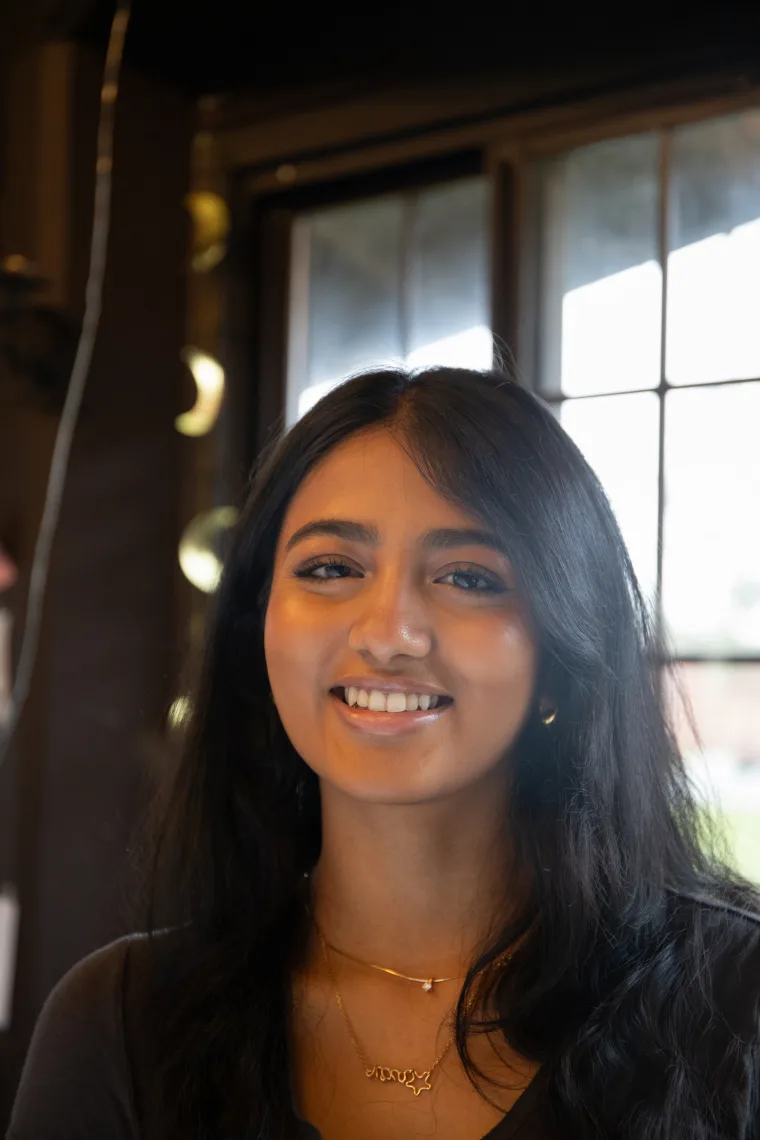 Person smiling indoors with long dark hair, wearing gold jewelry. Background features a window and dark walls.