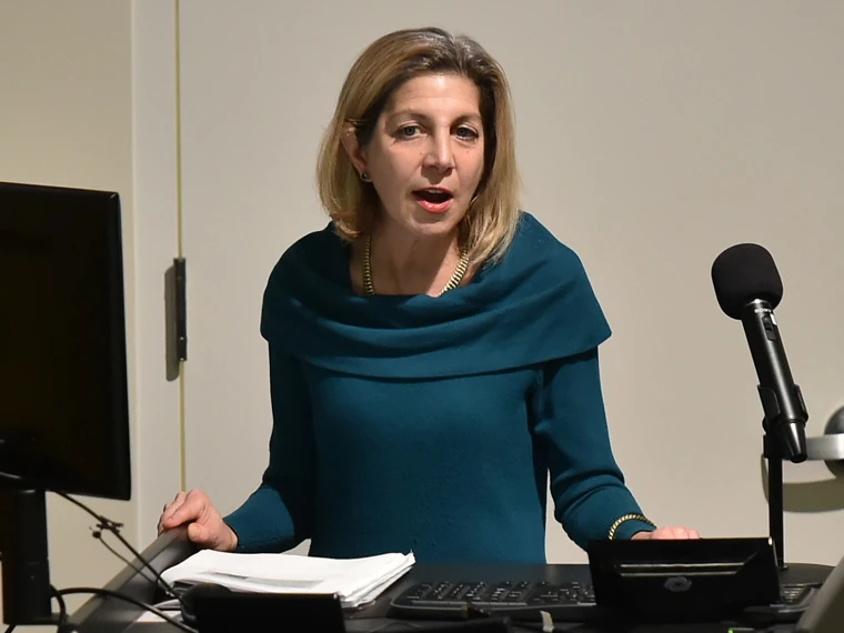 Person in a teal top speaking at a podium, with a microphone and computer nearby.