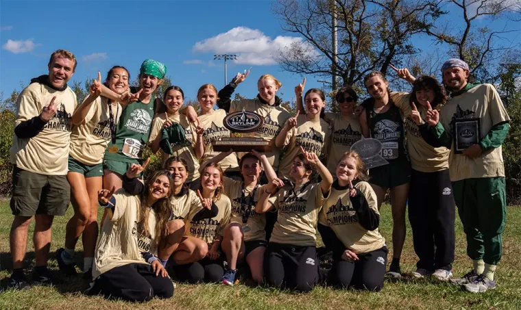 Group of people wearing matching championship t-shirts, smiling and holding a large trophy outdoors on a sunny day.