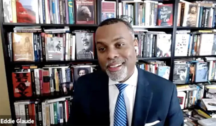 Individual wearing a suit and tie is smiling in front of a full bookshelf. A name label reads "Eddie Glaude" in the corner.