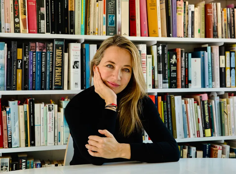 Person sitting at a table, resting head on hand, in front of a bookshelf filled with various books.