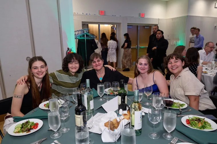Group of five smiling friends sitting around a dinner table with salads, wine bottles, and glasses. Others are in the background at the event.