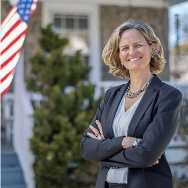 Person in a suit smiling with crossed arms in front of a house with a U.S. flag and greenery in the background.