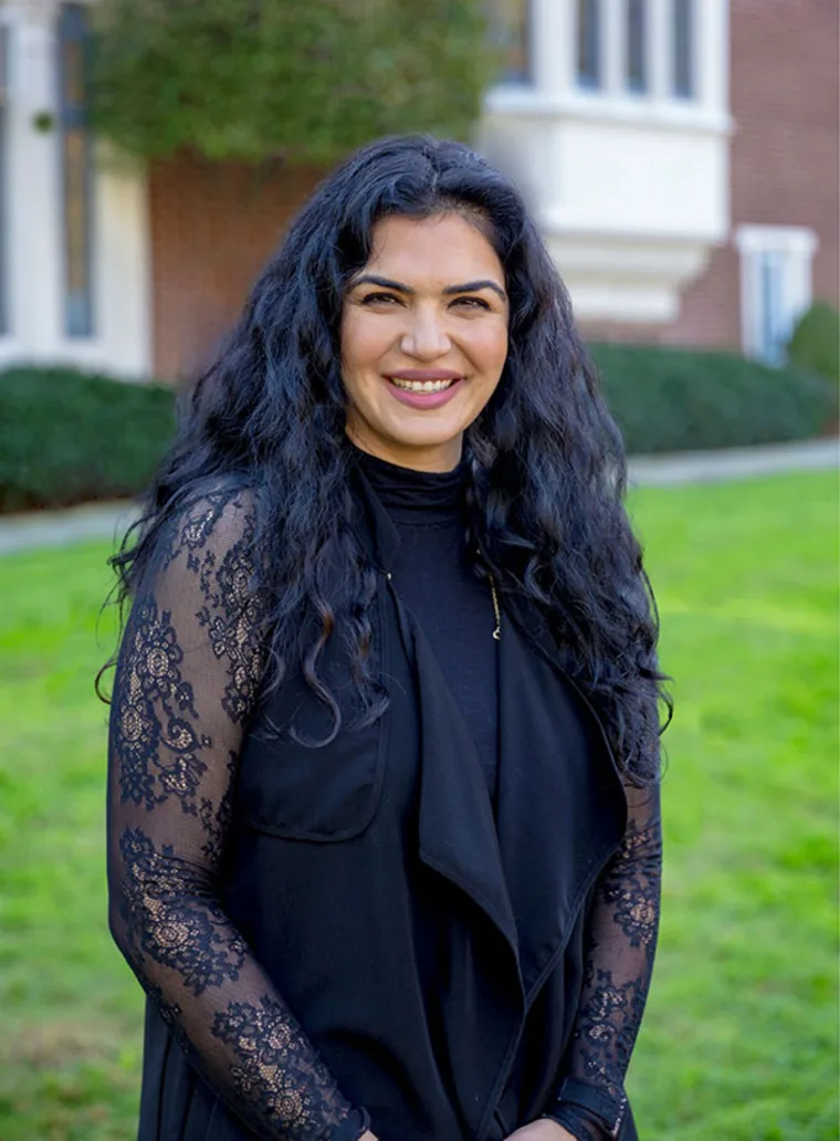 Person with long curly hair smiles while standing on grass in front of a brick building. Wearing a black outfit with lace sleeves.