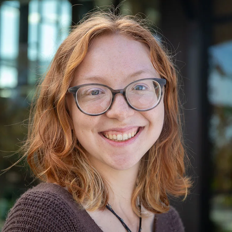 Person with red hair and glasses smiling, wearing a brown sweater, standing outdoors with blurred background.