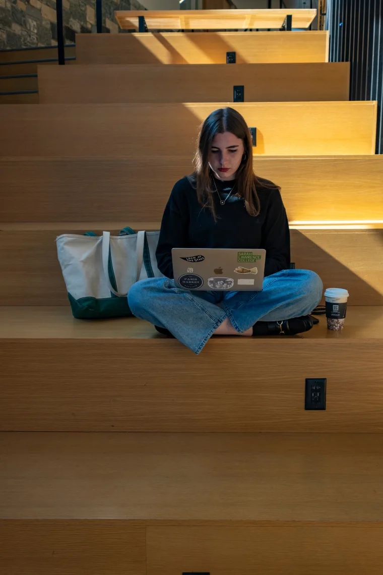 Person sitting on wooden steps, focused on a laptop. A large tote bag and a cup of coffee are nearby. Sunlight creates patterns on the steps.
