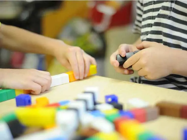 Two individuals assembling colorful interlocking blocks on a table. One person wears a striped shirt.