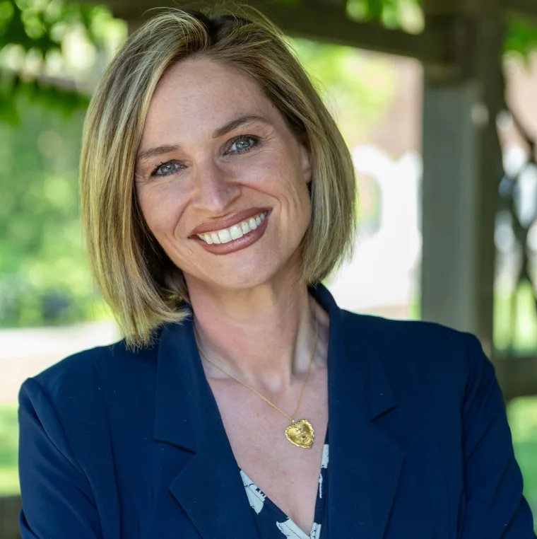 Person with short blond hair, smiling, wearing a blue blazer and heart-shaped necklace, standing outside with greenery in the background.