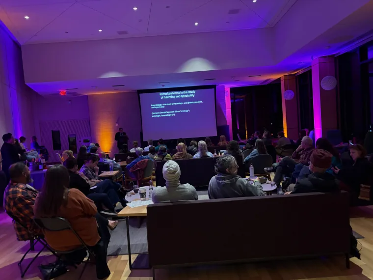 A group of people seated in a dimly lit room watch a presentation on a large screen. The room is illuminated with purple lighting.