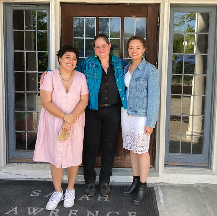 Three people standing together outside a building with glass doors and windows. The mat at their feet reads "Sarah Lawrence."