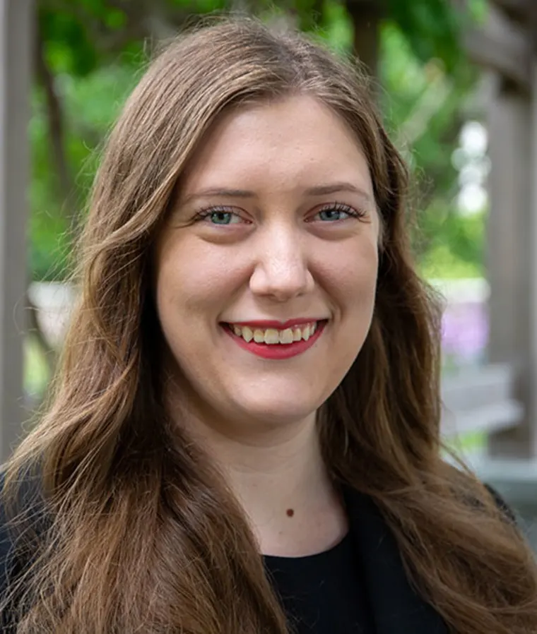 Person with long brown hair smiling outdoors, wearing a black top, with green foliage in the background.
